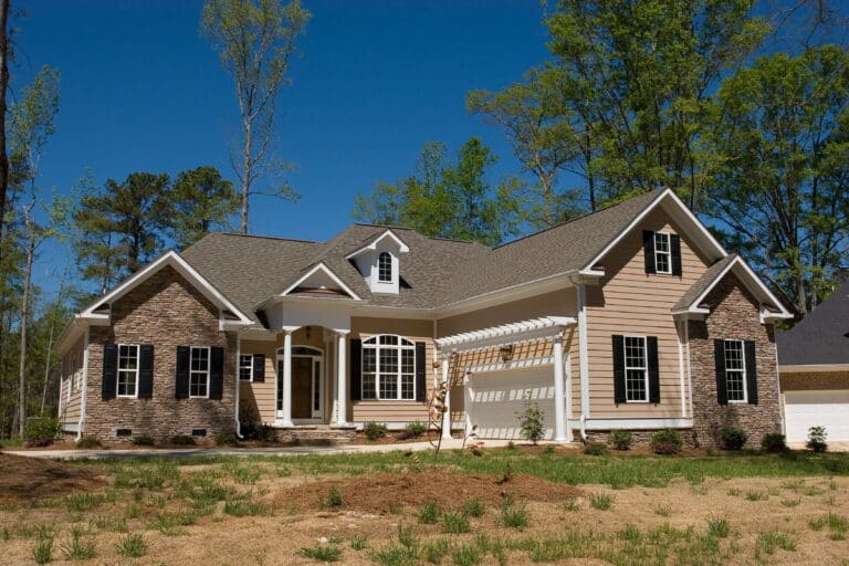 Single-story suburban house with stone and beige siding, white trim, gables, covered entry, pergola, sparse grass, trees, blue sky.