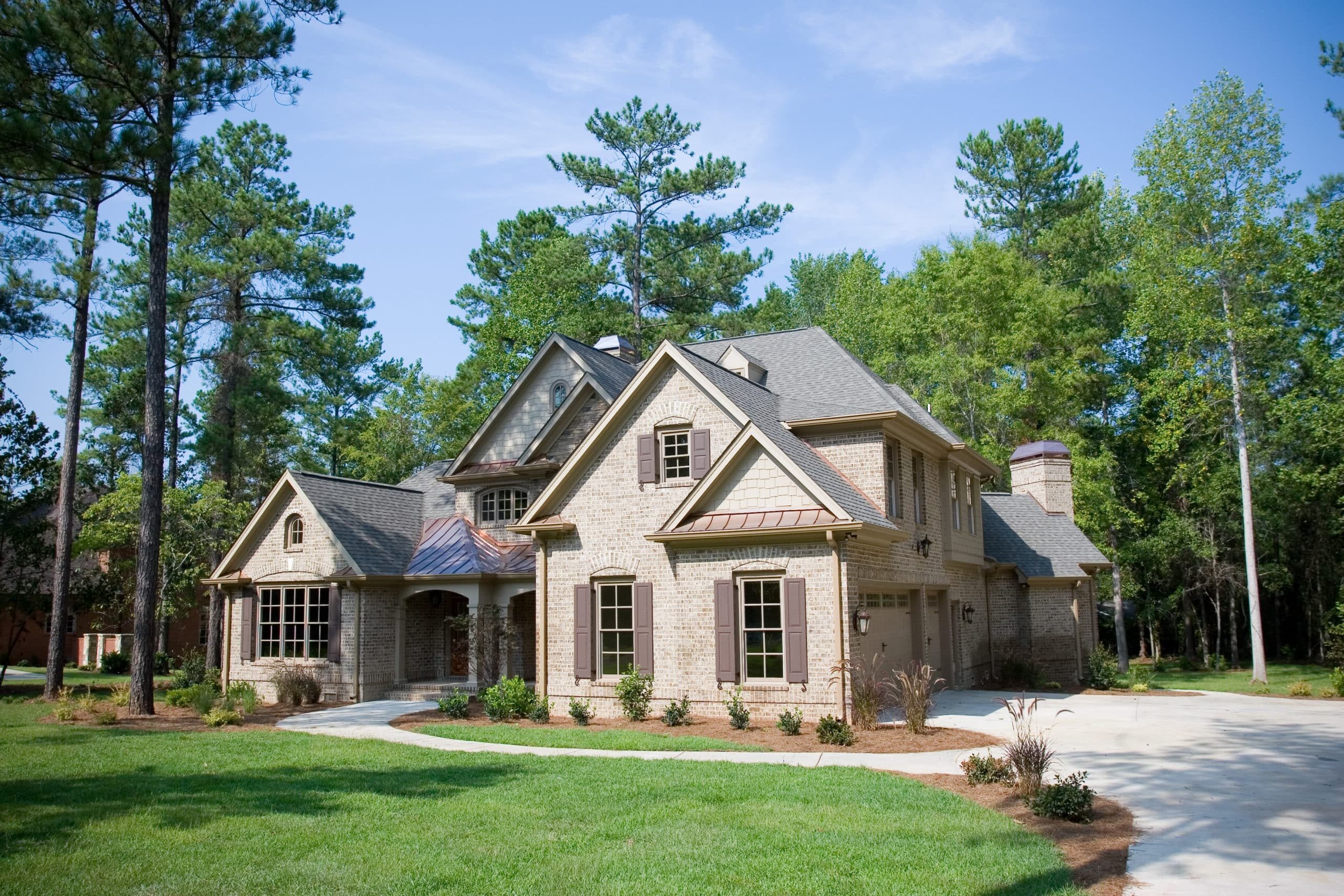 A large two-story brick house with peaked roofs, green lawn, tall trees, blue sky, wispy clouds, and a curved driveway to the garage.