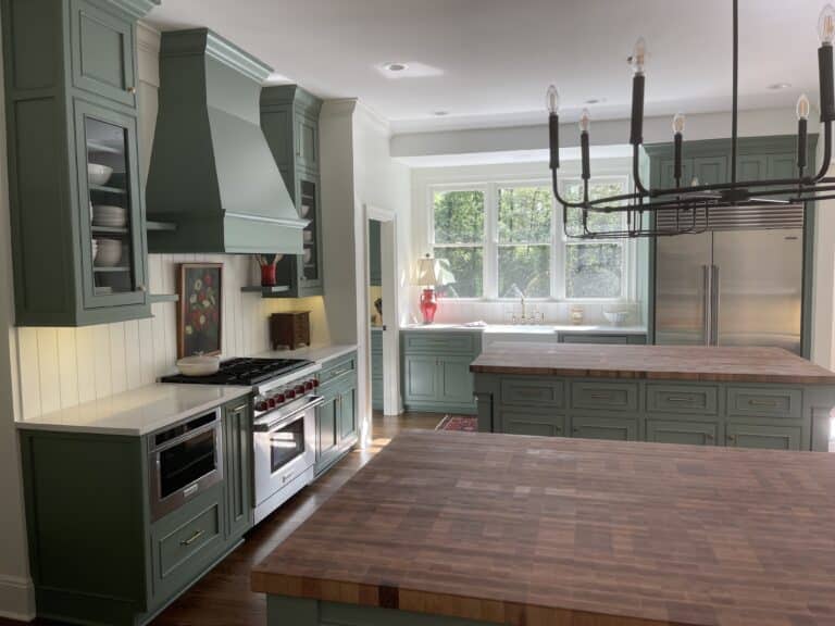 Spacious kitchen with green cabinets, butcher block counters, large stove, open shelves, farmhouse sink by three windows, black chandelier.