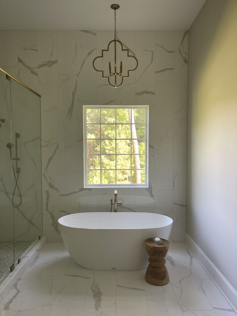 Modern bathroom featuring a white freestanding tub, wooden stool, marble surfaces, glass shower, big window, gold chandelier above.